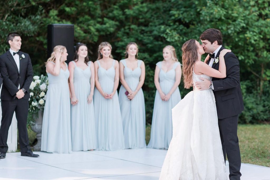 newlyweds share a kiss on dance floor during first dance while wedding party looks on