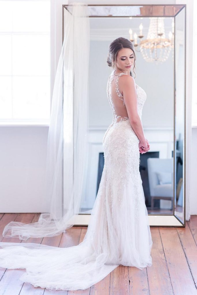 bride standing in front of mirror looking down shoulder with wedding veil draped over it