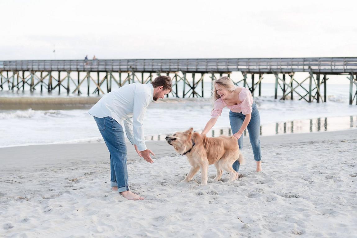 COuple playing with dog at beach with pier in the background