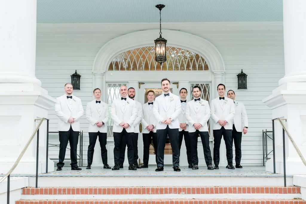 groom surrounded by groomsmen on front porch of house. All men dressed in white tuxedo jackets and black pants