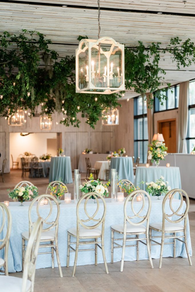 photo of wedding reception with greenery hanging from ceiling and table with blue linens with floral arrangement on top
