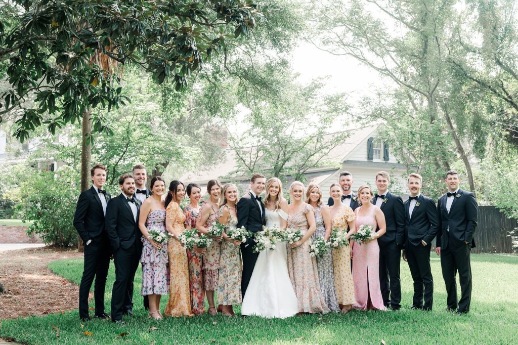 large wedding party in black tuxes and floral dresses stand in front yard with bride and groom