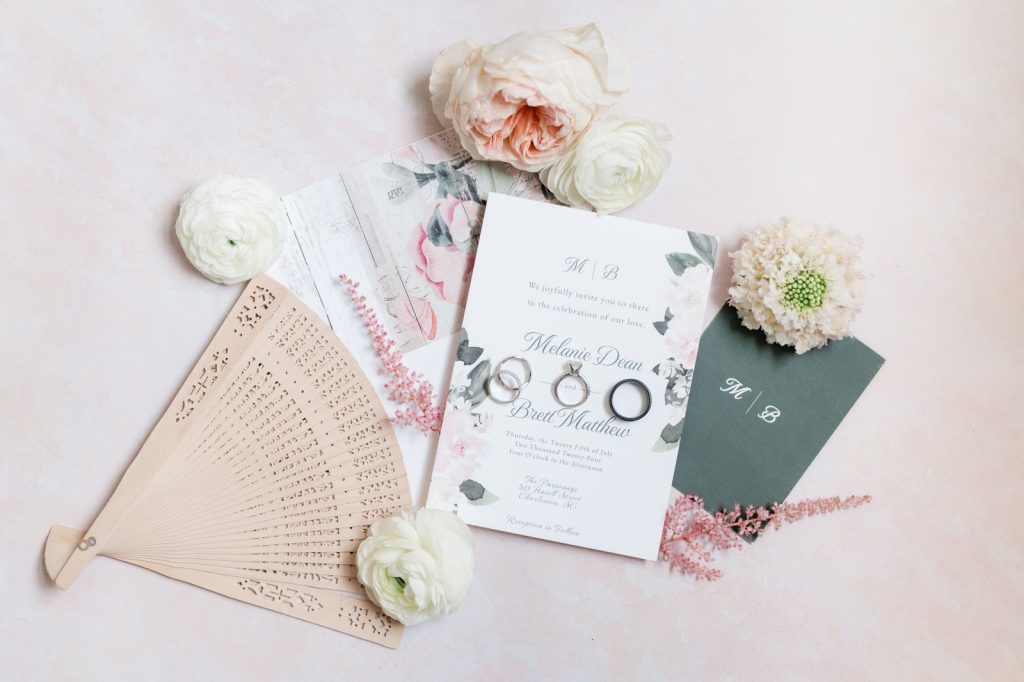 wedding invitation, rings, fan and flowers flat lay on a pink background