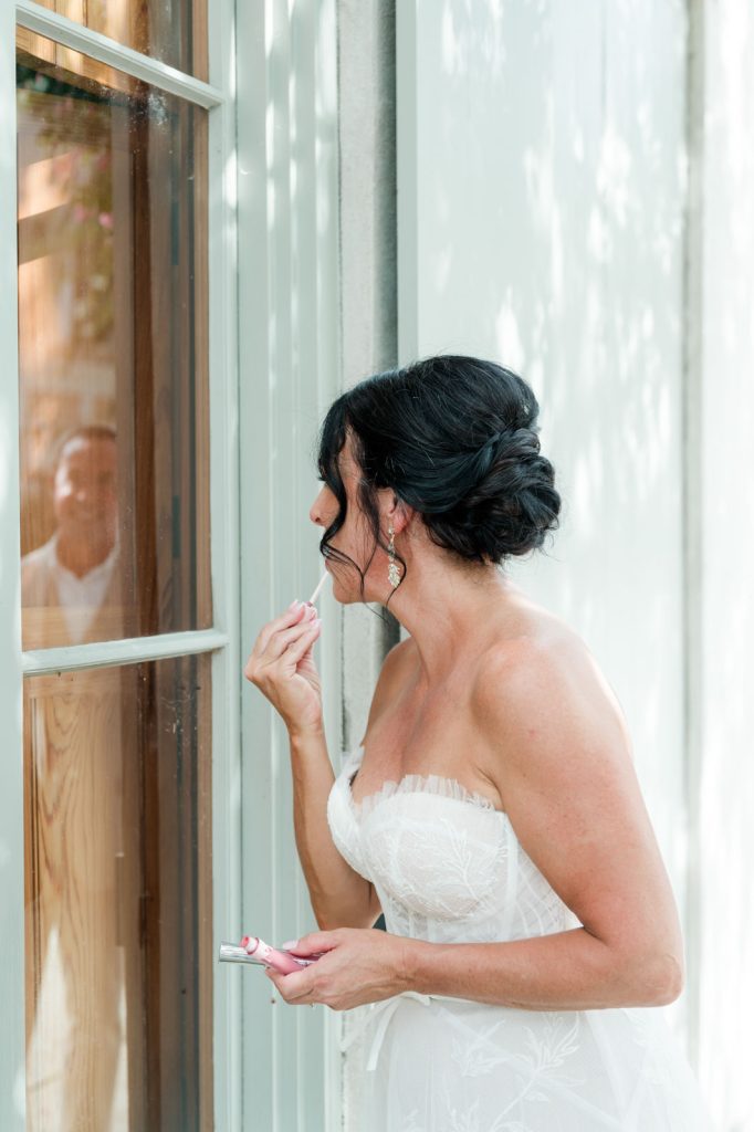 bride touches up lip gloss while looking at her reflection in a window. you can see the groom in the window's reflection