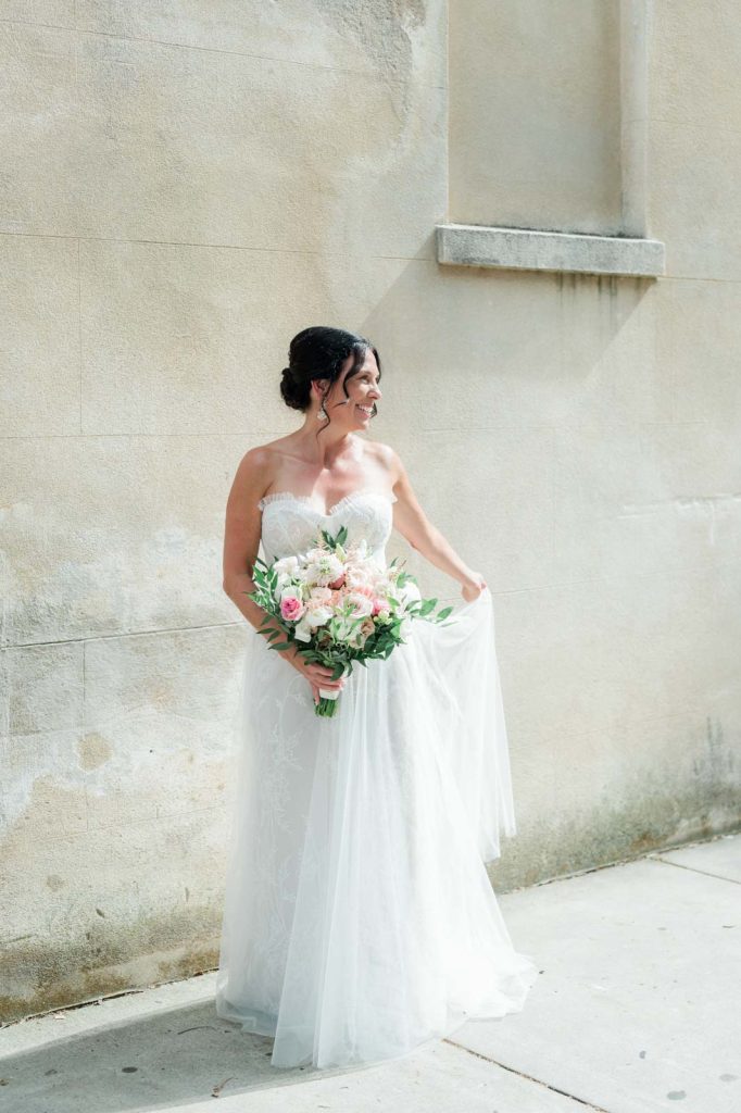 bride hold out wedding dress while holding floral bouquet and looking off camera at her groom
