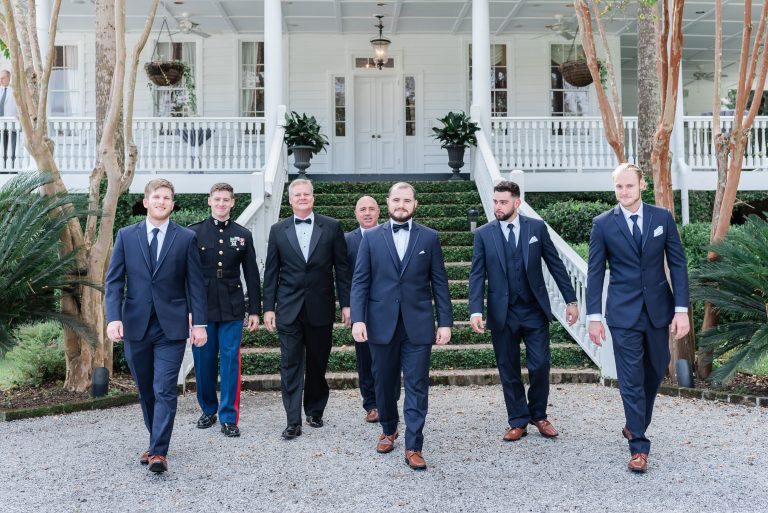Groom and groomsmen in navy blue tuxedos walk away from plantation house