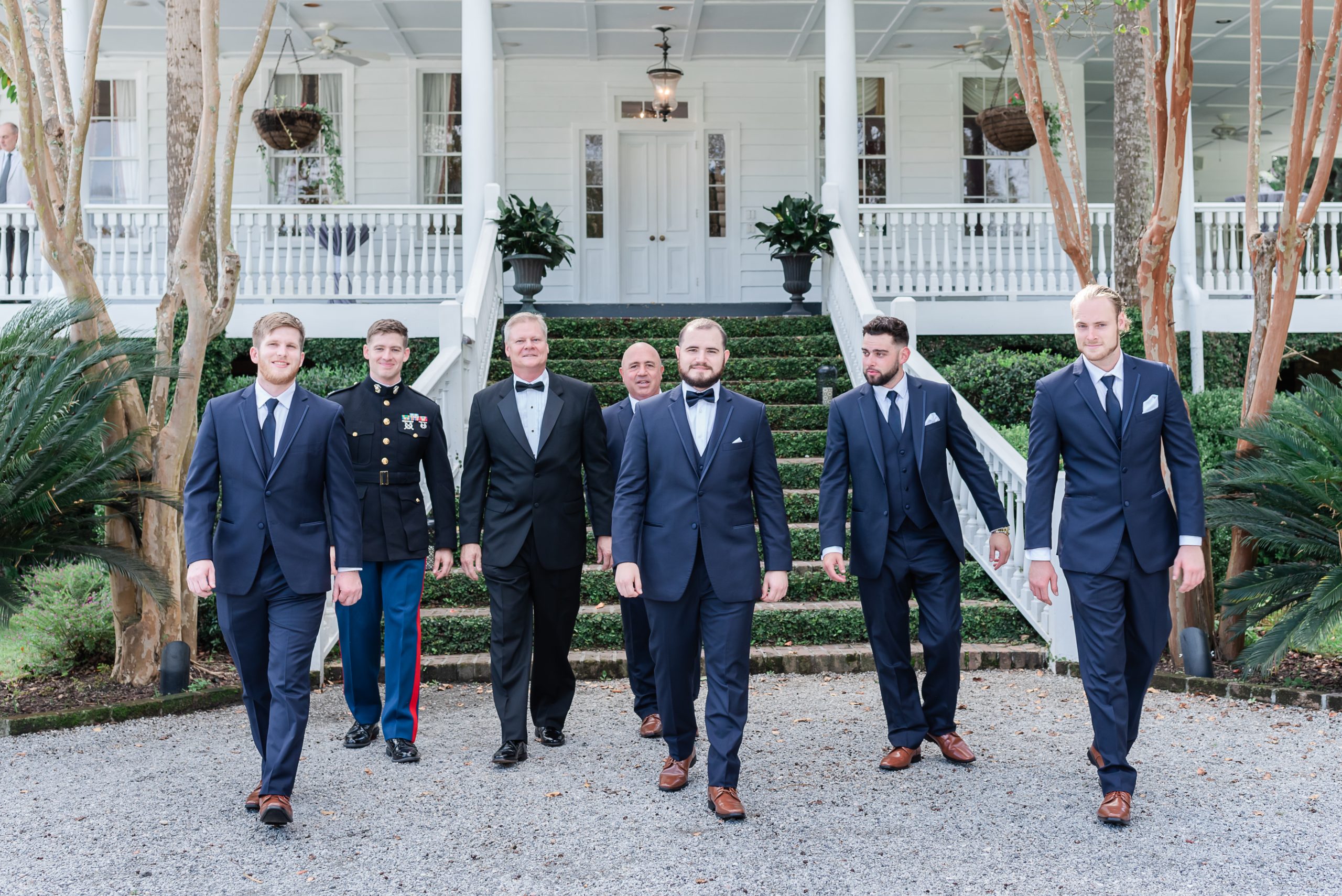 Groom and groomsmen in navy blue tuxedos walk away from plantation house
