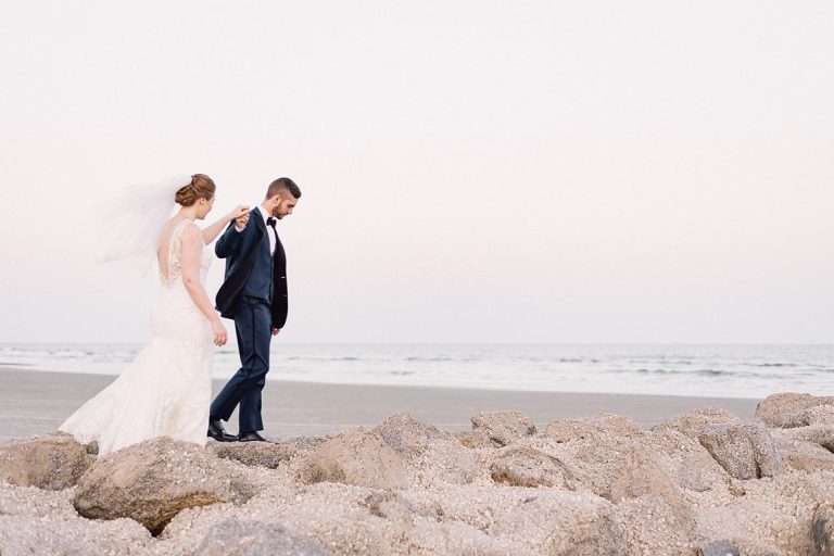 groom standing on rocks leading bride towards ocean