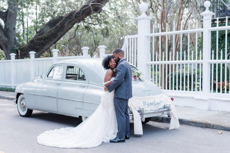 black newlywed couple embrace at the rear of their vintage gray get away car