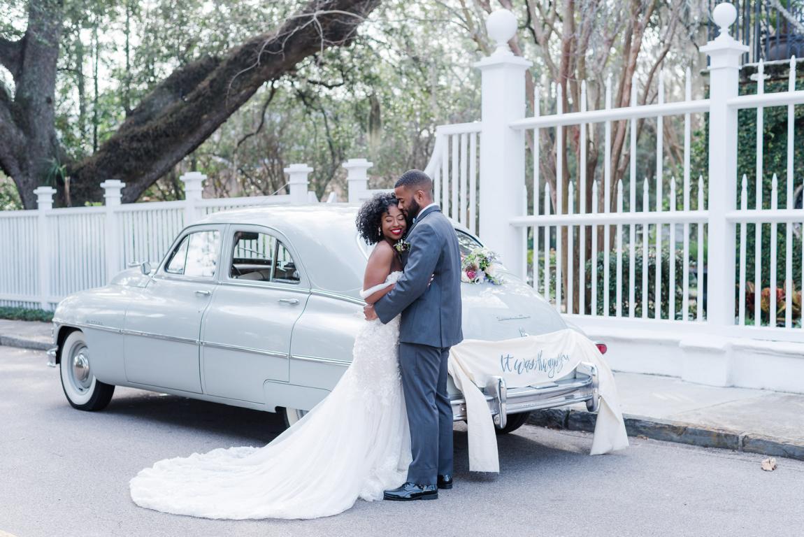 black newlywed couple embrace at the rear of their vintage gray get away car