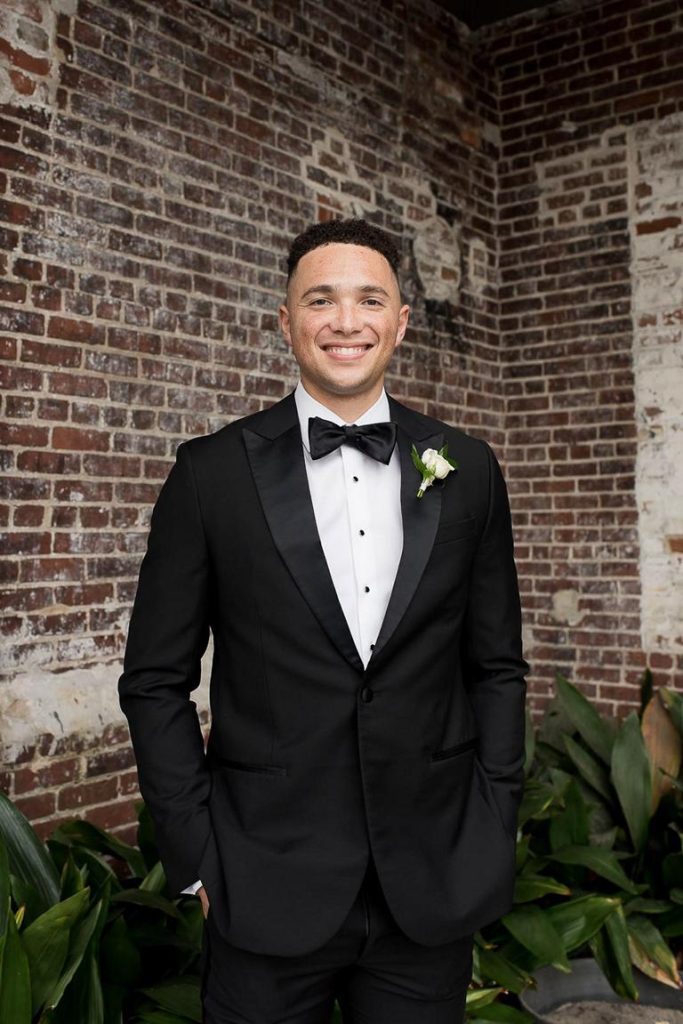 Groom in black tuxedo standing against brick building