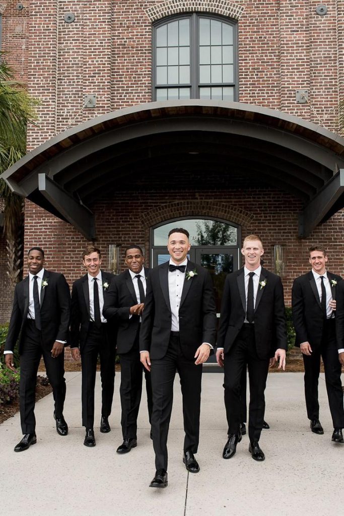 groom stands in the foreground with groomsmen behind him against brick building