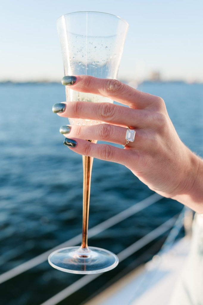 close up of hand holding a flute of champagne with an emerald cut engagement ring on over water
