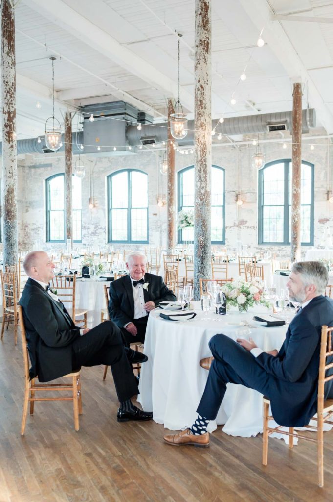 three men in tuxedos sit at wedding reception table before wedding