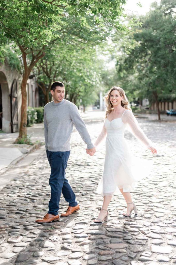 engaged couple walks across cobblestone street holding hands