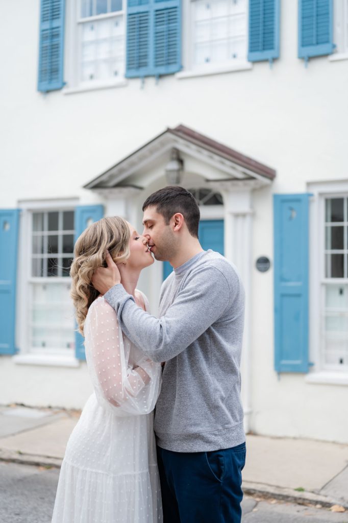 engaged couple kiss in front of white house with blue shutters